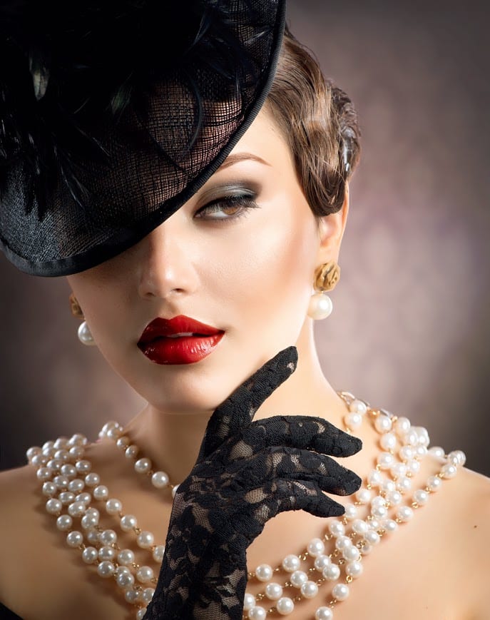 A portrait of a person wearing a Parisian hat, gloves, pear necklace, and red lipstick